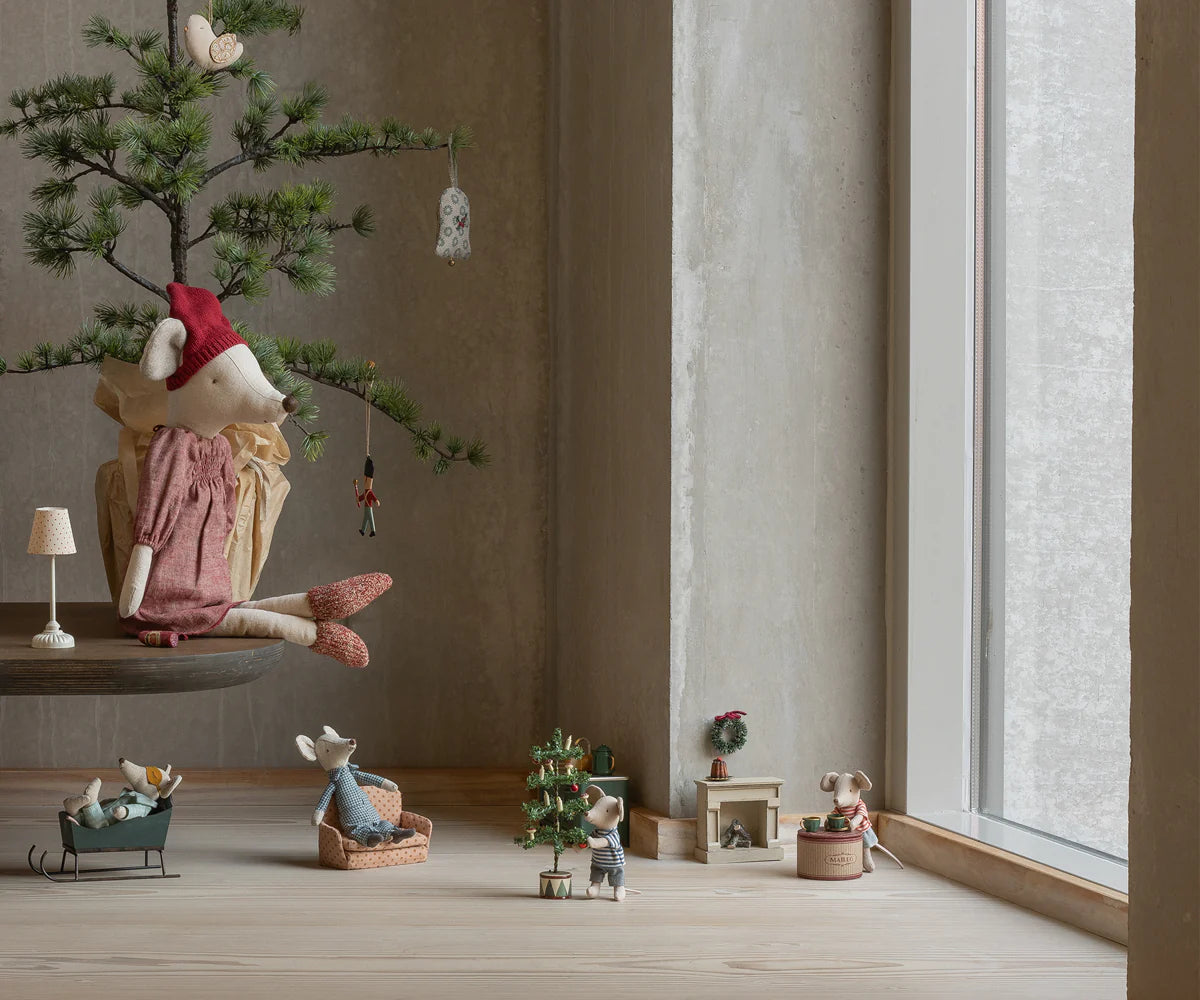 Maileg mouse dolls create a cozy scene: one in a red dress sits on a pine-needle branch while others decorate the Maileg Christmas Tree and play by a large window, all in a minimalist room.