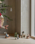Maileg mouse dolls create a cozy scene: one in a red dress sits on a pine-needle branch while others decorate the Maileg Christmas Tree and play by a large window, all in a minimalist room.