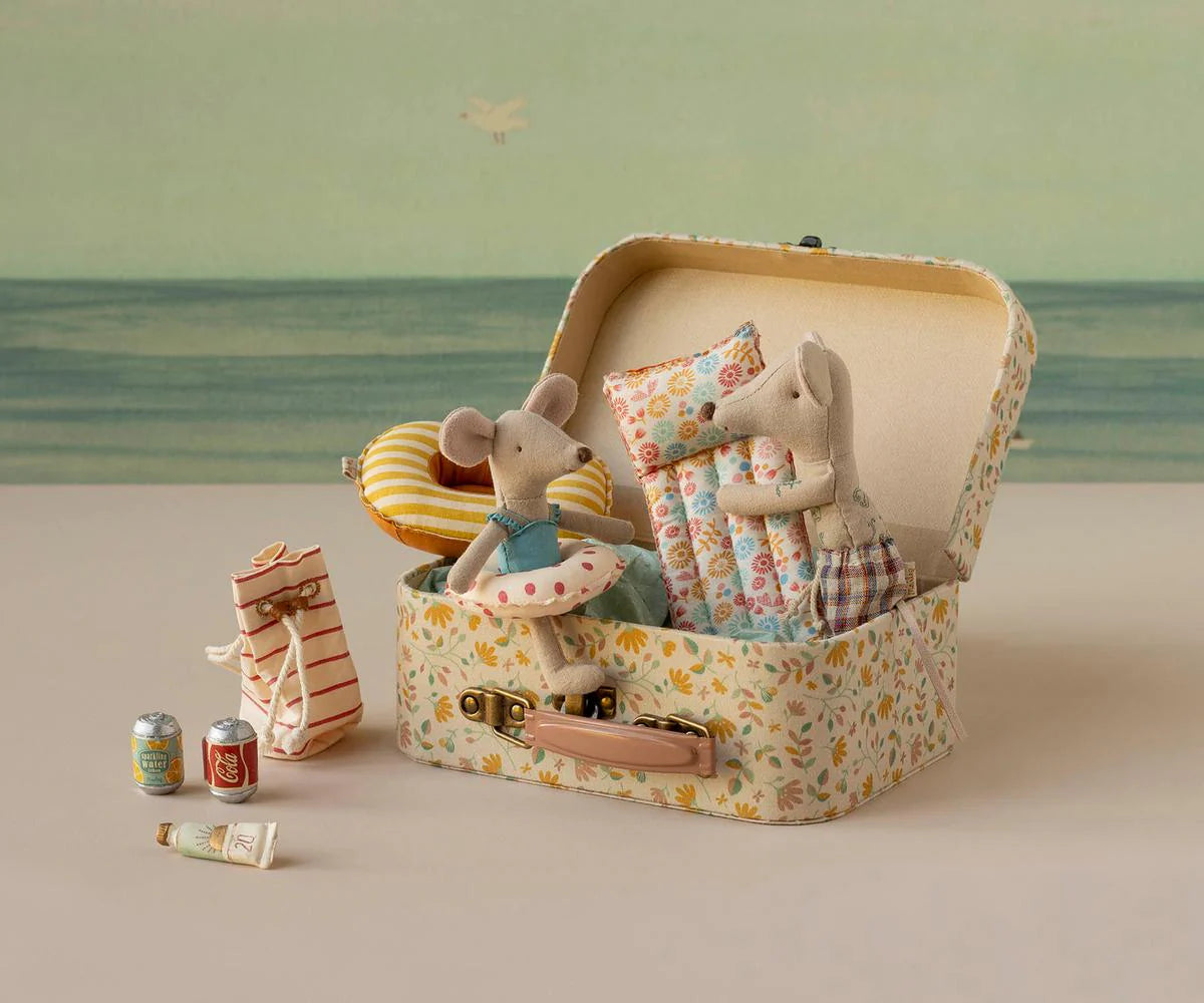 Maileg's Bag W/ Beach Essentials features two small mice in fabric clothes, sitting in a floral suitcase with pillows. Nearby are mouse-sized beach accessories: cans, tube, bag, striped float, and a seaside background for perfect play.