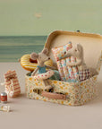 Maileg's Bag W/ Beach Essentials features two small mice in fabric clothes, sitting in a floral suitcase with pillows. Nearby are mouse-sized beach accessories: cans, tube, bag, striped float, and a seaside background for perfect play.