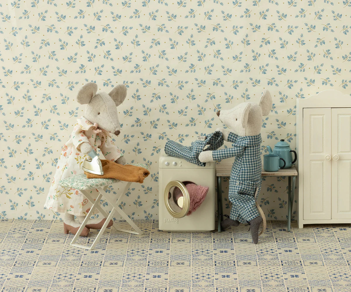 Two Maileg mice from the mouse family bustle in a dollhouse laundry room, using the Maileg Washing Machine. One mouse irons clothes while another loads laundry, with cozy accessories like a wardrobe and table set against floral wallpaper.
