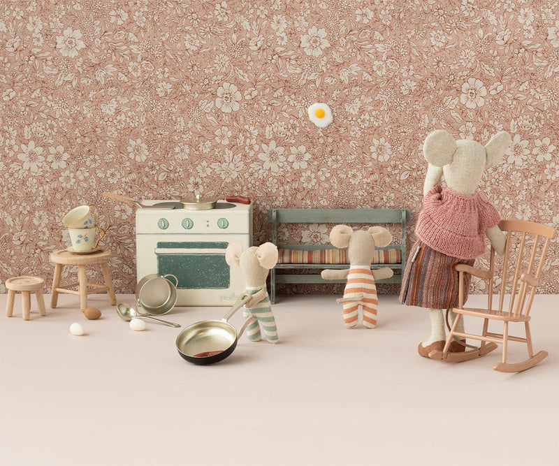 The Maileg Triplet Mice by Maileg snuggle in a tiny, vintage-themed kitchen with a stove, rocking chair, pans, and eggs; one mouse stands on a chair flipping a fried egg while the others look on.