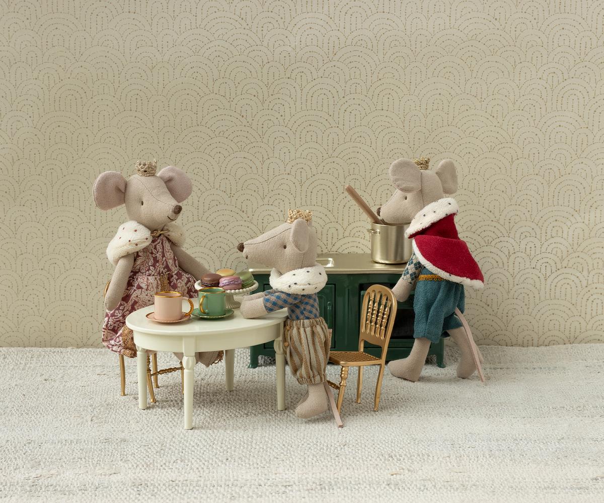 Three Maileg mice in royal outfits gather for tea at a tiny table with cups and pastries. One stirs a pot on a green stove, while the Maileg Gold Chair by Maileg adds charm to this whimsical, cozy tea party scene.