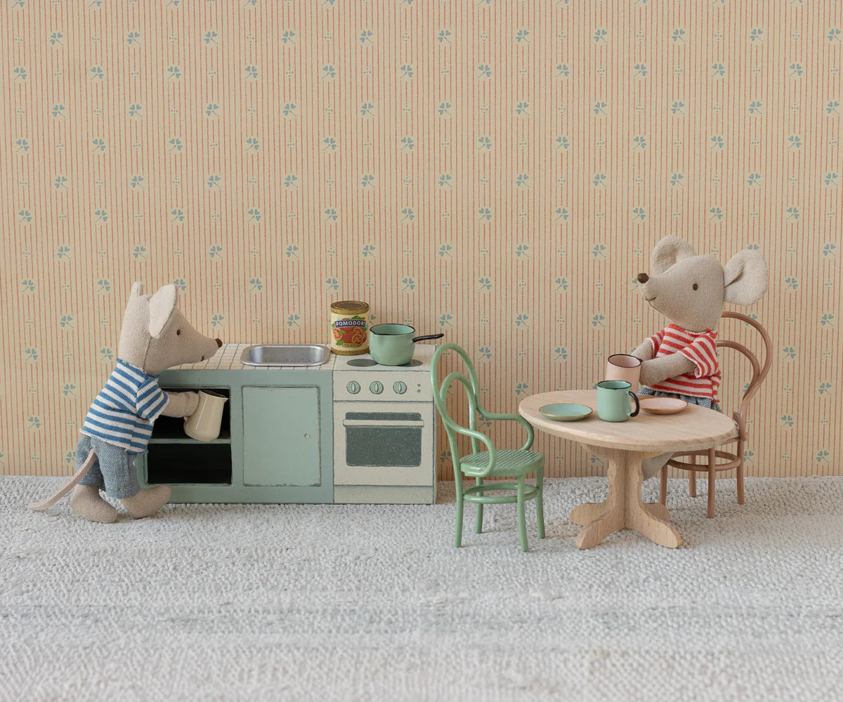 Two Maileg toy mice in striped shirts are in a miniature kitchen. One stands by the stove and sink, while the other sits at a table with cups and a plate on the Maileg Vintage Chair, surrounded by charming Maileg dollhouse accessories.