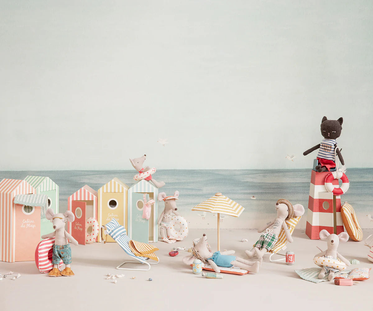 Maileg's Little Brother and friends don beach attire to relax and play by colorful cabanas. A stuffed cat perches nearby, all set for imaginative adventures with the Maileg Little Brother /Beach Cabana set.