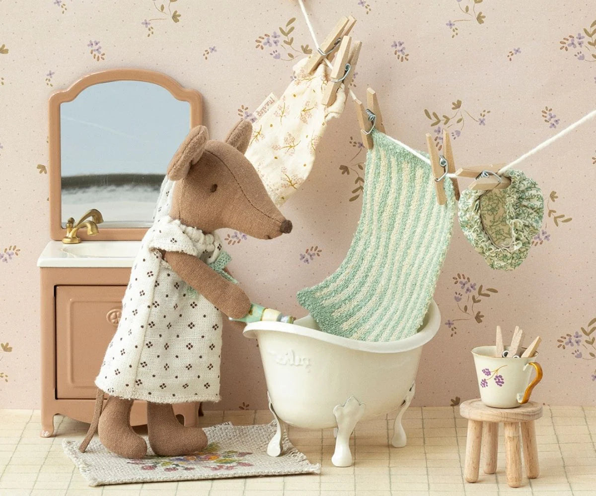 A brown fabric mouse in a dress washes clothes beside the Maileg Bathtub. Clothes hang above, with a sink, mirror, and stool with clothespins adding charm to this dollhouse scene featuring Maileg accessories.