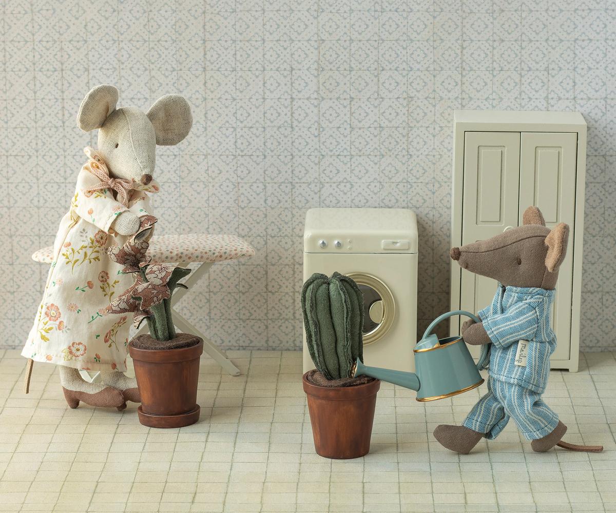Two toy mice in pajamas stand in a laundry room with patterned wallpaper and tiled floor. One mouse irons, while the other waters a green plant in the Maileg Flower Pot by Maileg. An ironing board, washing machine, and cabinet are nearby.