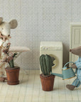 Two toy mice in pajamas stand in a laundry room with patterned wallpaper and tiled floor. One mouse irons, while the other waters a green plant in the Maileg Flower Pot by Maileg. An ironing board, washing machine, and cabinet are nearby.
