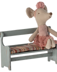 A Maileg stuffed mouse in a pink dress and headband sits atop the Maileg Bench, which features a striped cushion for comfort on its small, rustic wooden seat.
