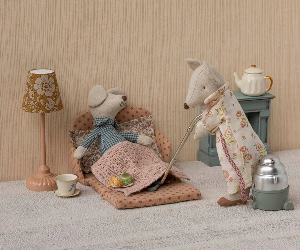 Two Maileg stuffed mice relax in a cozy living room: one naps under a blanket on a polka-dot bed while the other, in an apron, vacuums by the Maileg Sofa Bed. A lamp, teacup, and teapot complete this inviting Maileg brand scene.