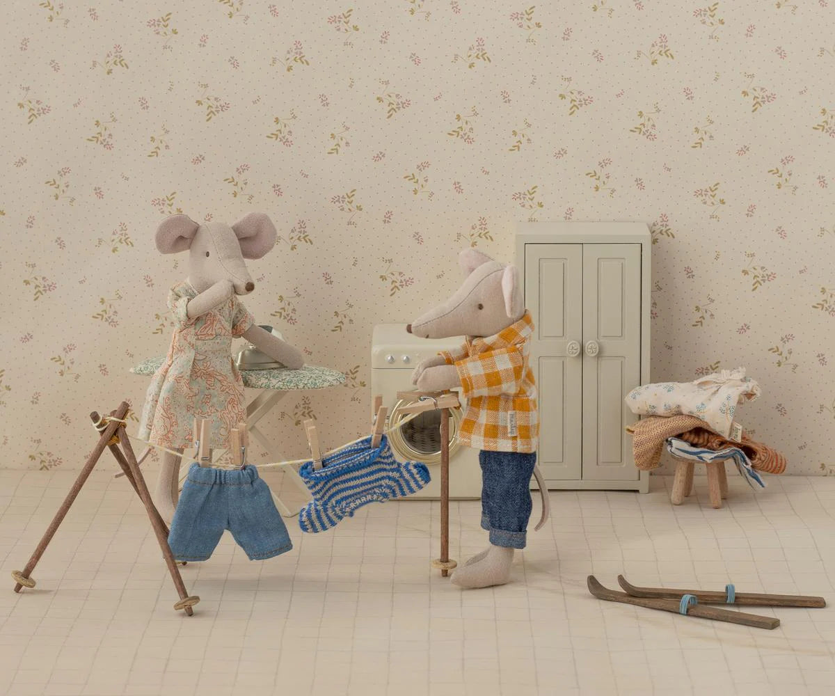 Maileg Dad Mouse wears a plaid shirt and helps hang tiny clothes on a drying rack in his dollhouse room, which features Maileg accessories like a small wardrobe, folded laundry on a stool, and toy skis on the floor.