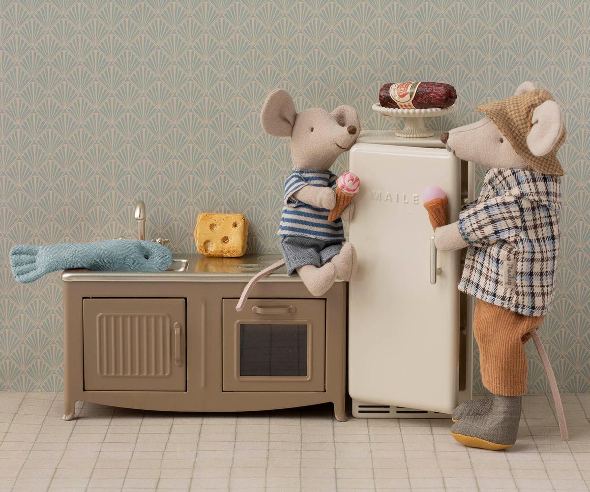 Two Maileg toy mice in clothes enjoy ice cream by the Maileg Kitchen set, which features a fridge topped with a fish, cheese, and pastry. Patterned wallpaper in the background adds extra charm to this cozy scene.