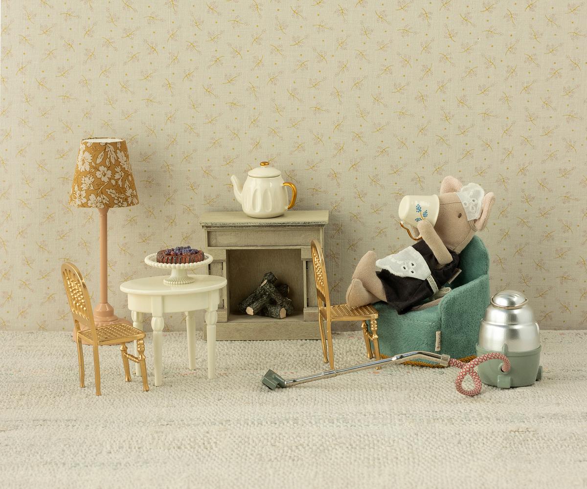 Maileg’s Big Sister Maid Mouse relaxes in an armchair, sipping tea in a miniature Maileg living room with a table, chairs, lamp, fireplace, teapot, pie, and a vacuum cleaner on the floor.