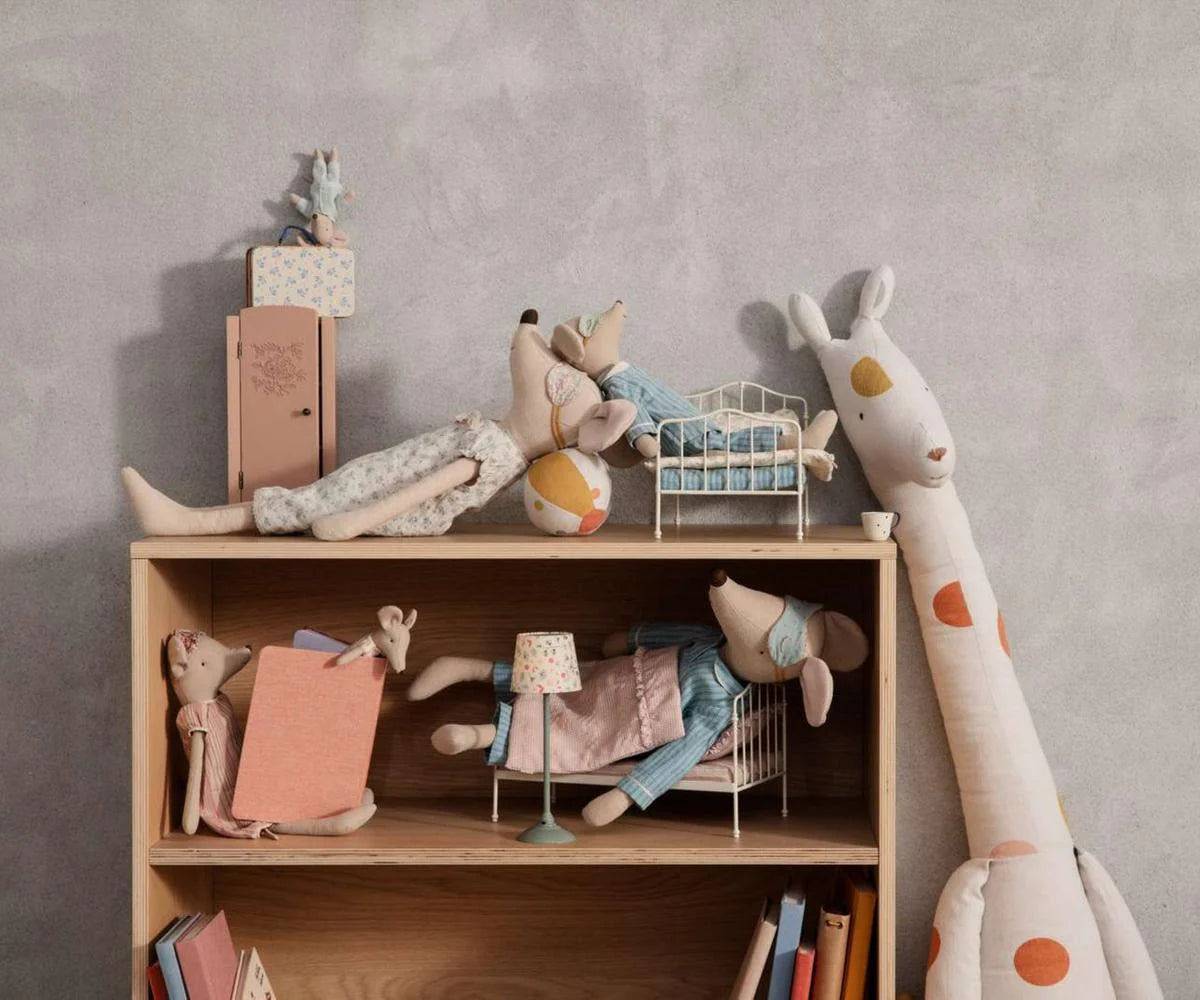 A wooden shelf shows Maileg plush toys and furniture, featuring the Maileg Medium Mouse in a nightgown as a cozy bedtime friend, with small toy beds, miniature furniture, books, and a tall stuffed giraffe against a plain light gray wall.
