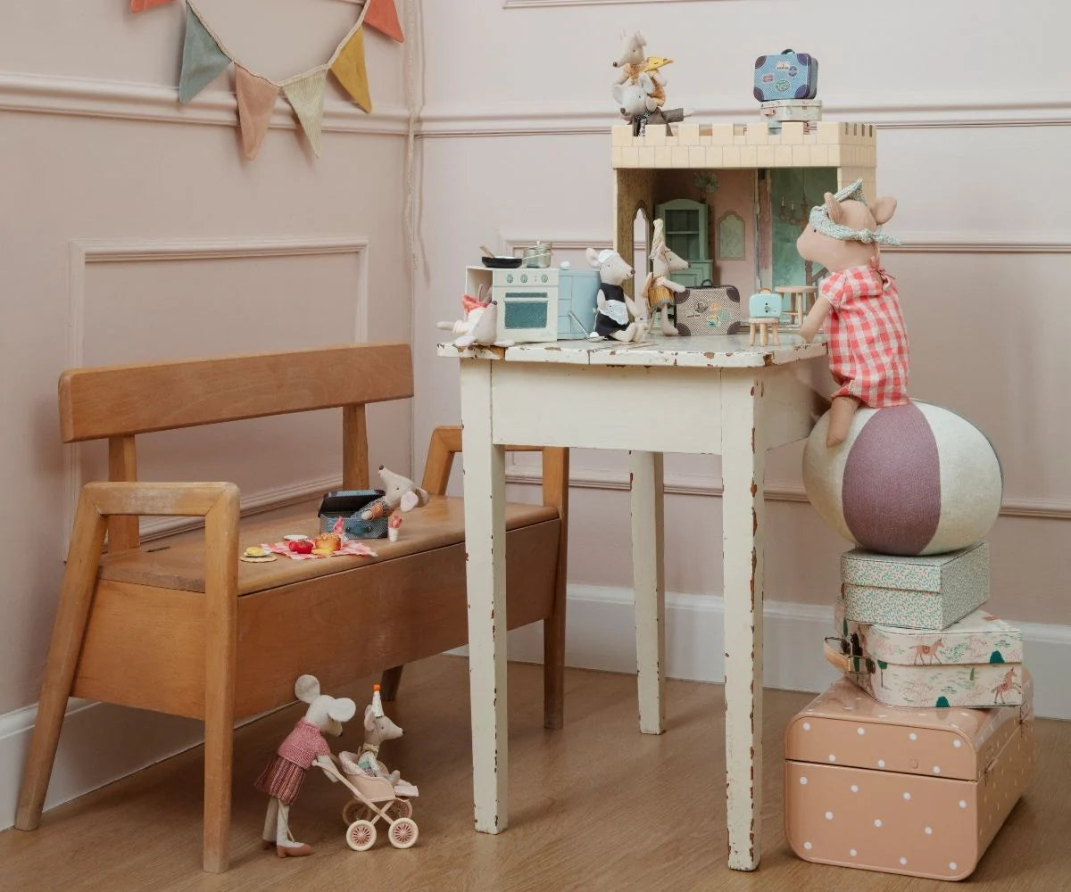A cozy playroom features a wooden bench, a Maileg toy mouse in a stroller, and a white table with the Maileg Castle Hall dollhouse. One mouse stands on boxes and a ball, ready for magical tea parties and royal adventures by Maileg.