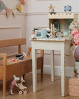 A cozy playroom features a wooden bench, a Maileg toy mouse in a stroller, and a white table with the Maileg Castle Hall dollhouse. One mouse stands on boxes and a ball, ready for magical tea parties and royal adventures by Maileg.