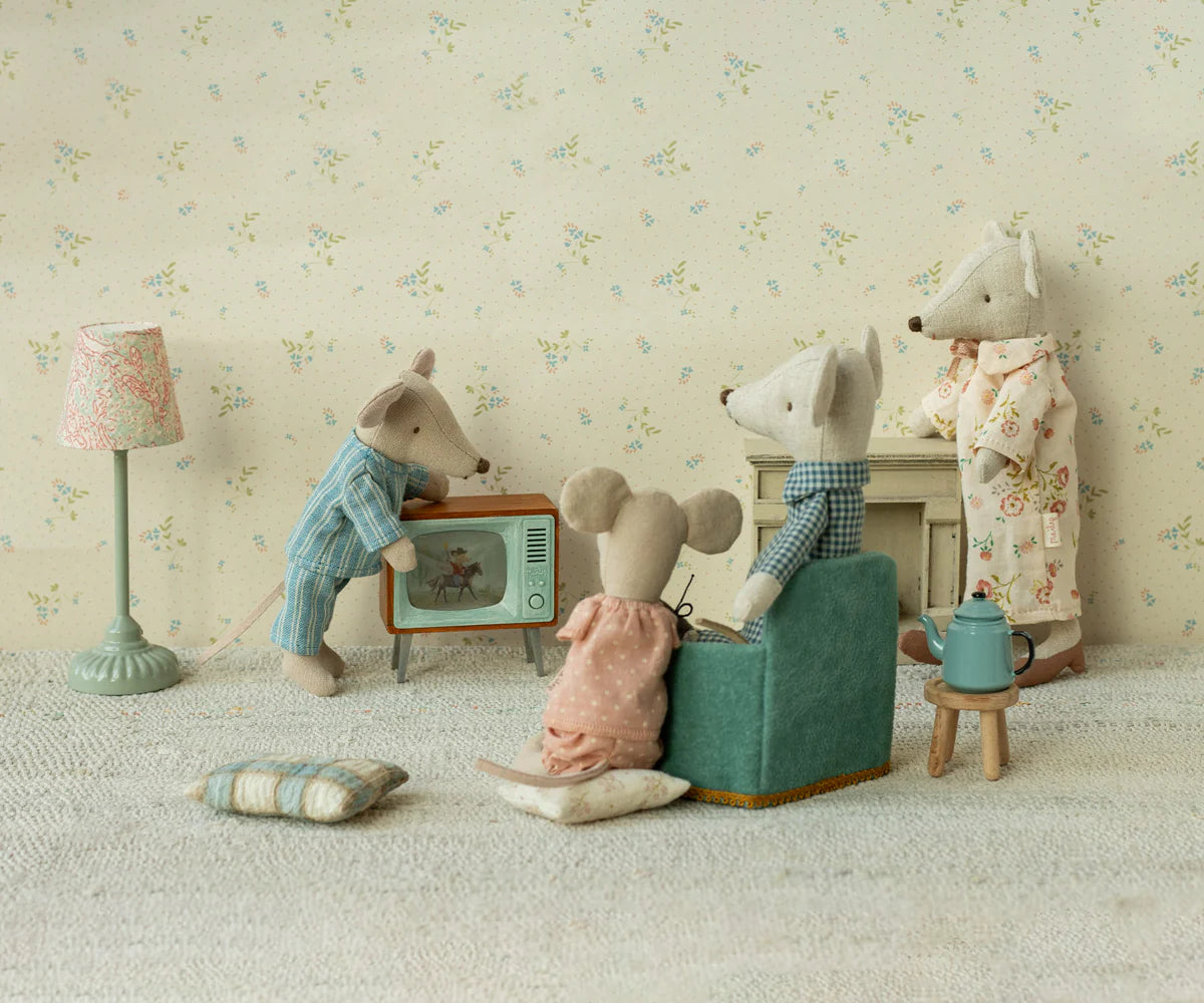 Four Maileg mice in pajamas relax in their cozy mouse home featuring the Maileg Television. Two lounge on a sofa, one adjusts the TV, and another stands by the fireplace. Wooden decor and toy accessories create a charming living room by Maileg.