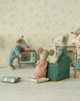Four Maileg mice in pajamas relax in their cozy mouse home featuring the Maileg Television. Two lounge on a sofa, one adjusts the TV, and another stands by the fireplace. Wooden decor and toy accessories create a charming living room by Maileg.