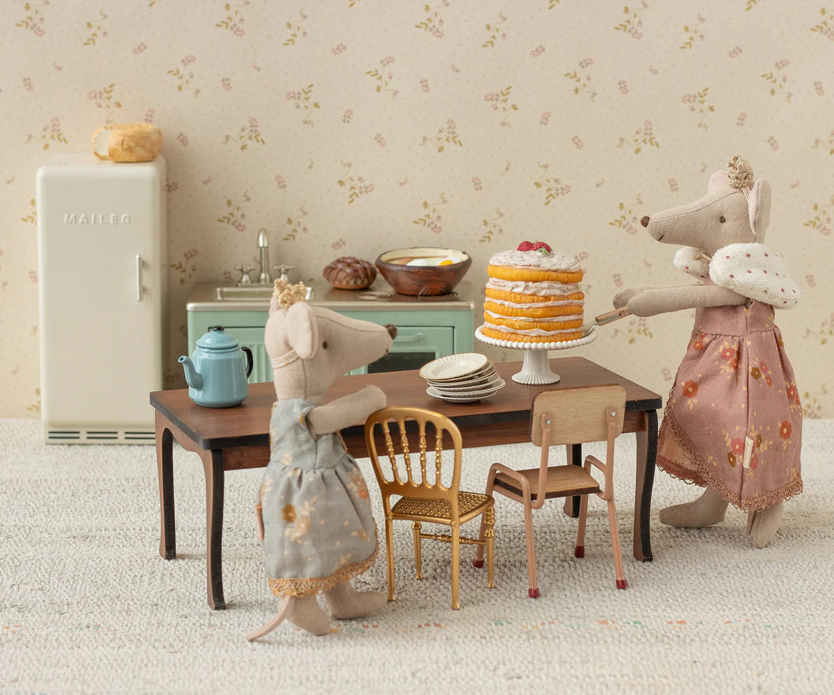 Two Maileg mice in dresses stand in a vintage kitchen, with one serving cake on the Maileg Long Dining Table set with plates and a teapot. Dollhouse furniture and a retro fridge finish the charming scene.