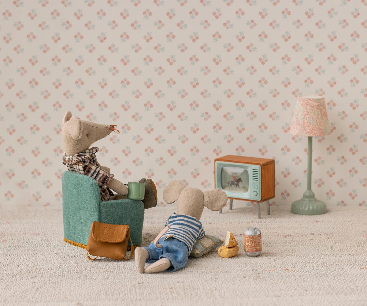 Two stuffed toy mice from Maileg relax in a cozy room featuring the Maileg Television. One mouse sits in a chair while the other lounges on the floor amid snacks, drinks, and accessories, with floral wallpaper enhancing the scene.