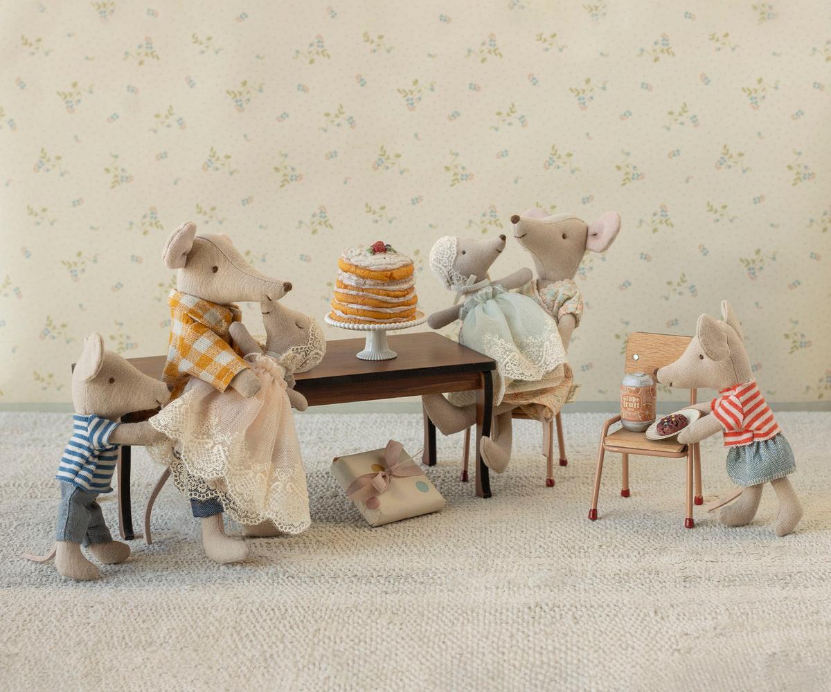 Five Maileg stuffed toy mice enjoy a stack of pancakes and a gift around the Maileg Long Dining Table. Two sit at the table, one cooks at a play stove, and two stand nearby, all in cute, detailed outfits. Brand: Maileg.