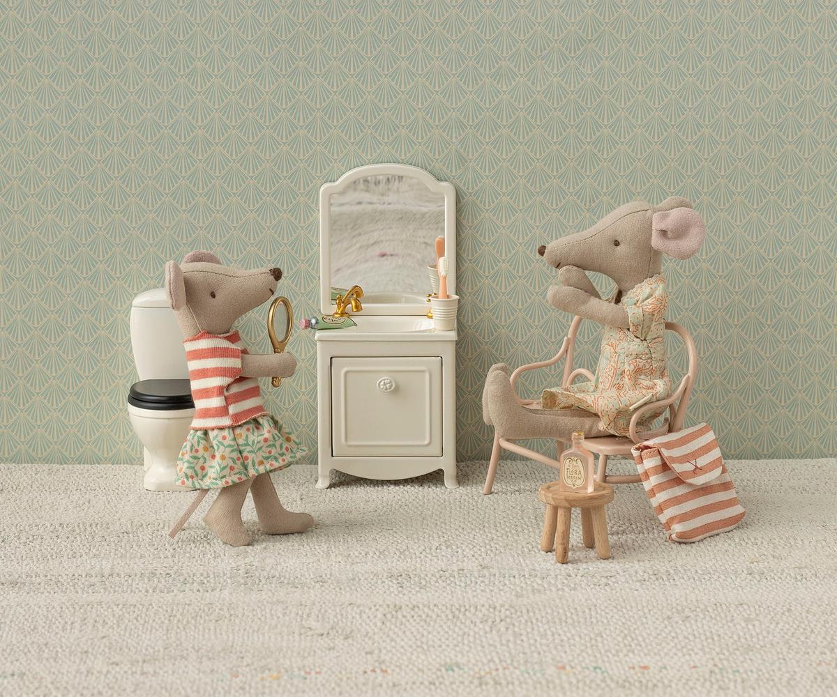 In a dollhouse bathroom, Maileg Mum Mouse by Maileg sits on a chair with a towel near a striped bag and accessories, while another mouse stands by the sink with a mirror; a toilet and vanity with toiletries are also present.
