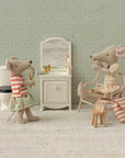 In a dollhouse bathroom, Maileg Mum Mouse by Maileg sits on a chair with a towel near a striped bag and accessories, while another mouse stands by the sink with a mirror; a toilet and vanity with toiletries are also present.