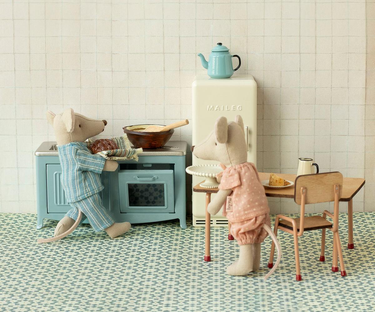 Two toy mice in pajamas are in a cozy kitchen; one bakes at a blue stove while the other stands by the Maileg Table and Chairs set. A vintage-style fridge and teapot complete this charming scene from Maileg.