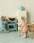 Two toy mice in pajamas are in a cozy kitchen; one bakes at a blue stove while the other stands by the Maileg Table and Chairs set. A vintage-style fridge and teapot complete this charming scene from Maileg.