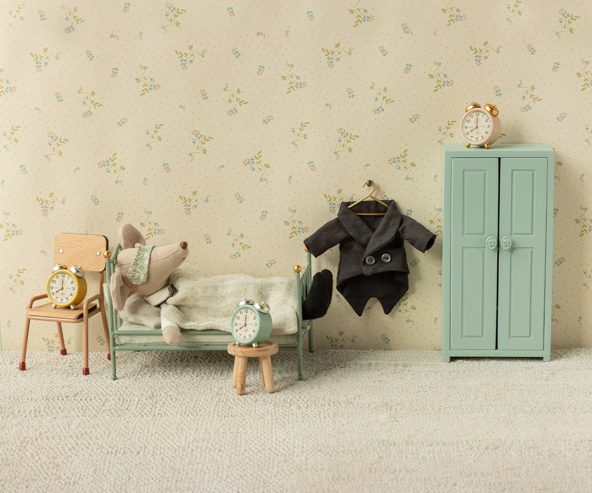 A small bedroom scene shows a Maileg mouse doll in bed with four Maileg Alarm Clocks, a metal chair, wardrobe, stool, and tiny suit on floral wallpaper.