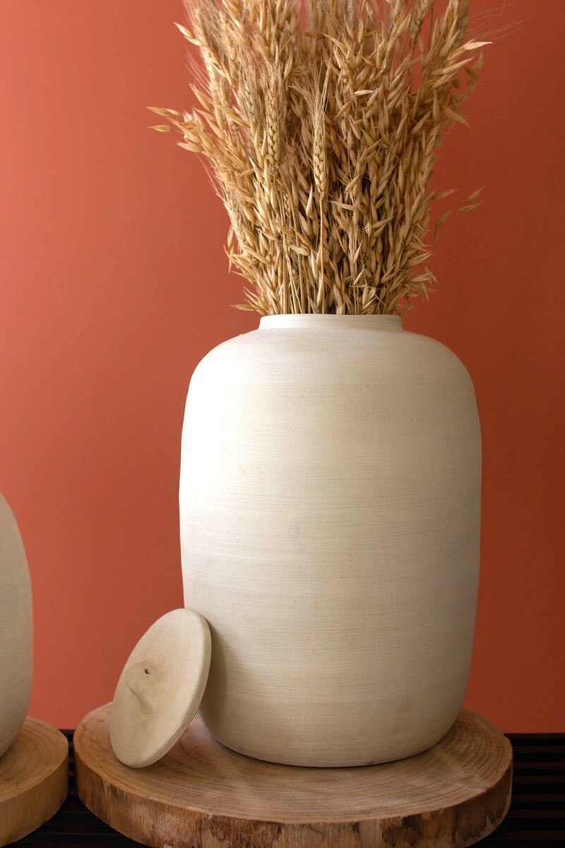 An Off-White Ceramic Canister by Kalalou, Inc sits on a round wooden base, its lid resting nearby. Filled with dried wheat, it evokes classic kitchen storage. The background is a warm solid orange.
