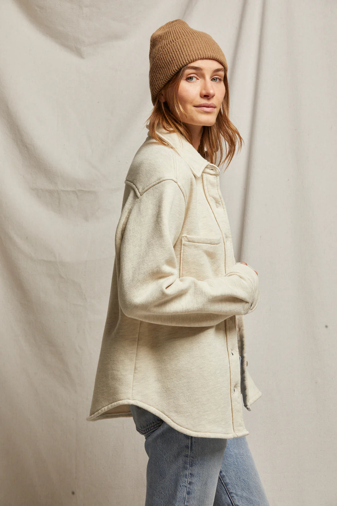 A person wearing the Cypress Fleece Button Down Jacket by Perfectwhitetee in light beige and a tan beanie stands sideways, looking at the camera. They have light brown hair and are also sporting light blue jeans. The backdrop is a neutral fabric.