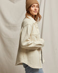 A person wearing the Cypress Fleece Button Down Jacket by Perfectwhitetee in light beige and a tan beanie stands sideways, looking at the camera. They have light brown hair and are also sporting light blue jeans. The backdrop is a neutral fabric.