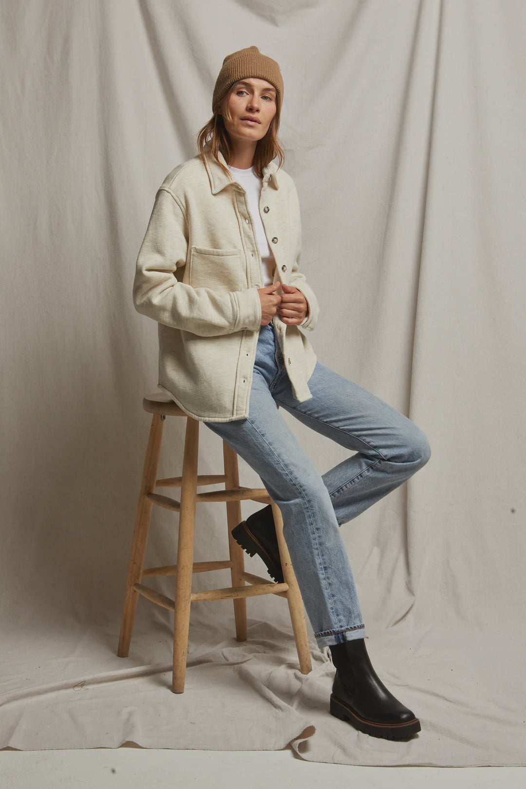 A person wearing the Cypress Fleece Button Down Jacket by Perfectwhitetee, paired with a white shirt, light blue jeans, black boots, and a brown beanie sits on a wooden stool against a neutral backdrop. They maintain a relaxed, casual pose with their hands holding the jacket.