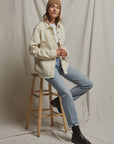 A person wearing the Cypress Fleece Button Down Jacket by Perfectwhitetee, paired with a white shirt, light blue jeans, black boots, and a brown beanie sits on a wooden stool against a neutral backdrop. They maintain a relaxed, casual pose with their hands holding the jacket.