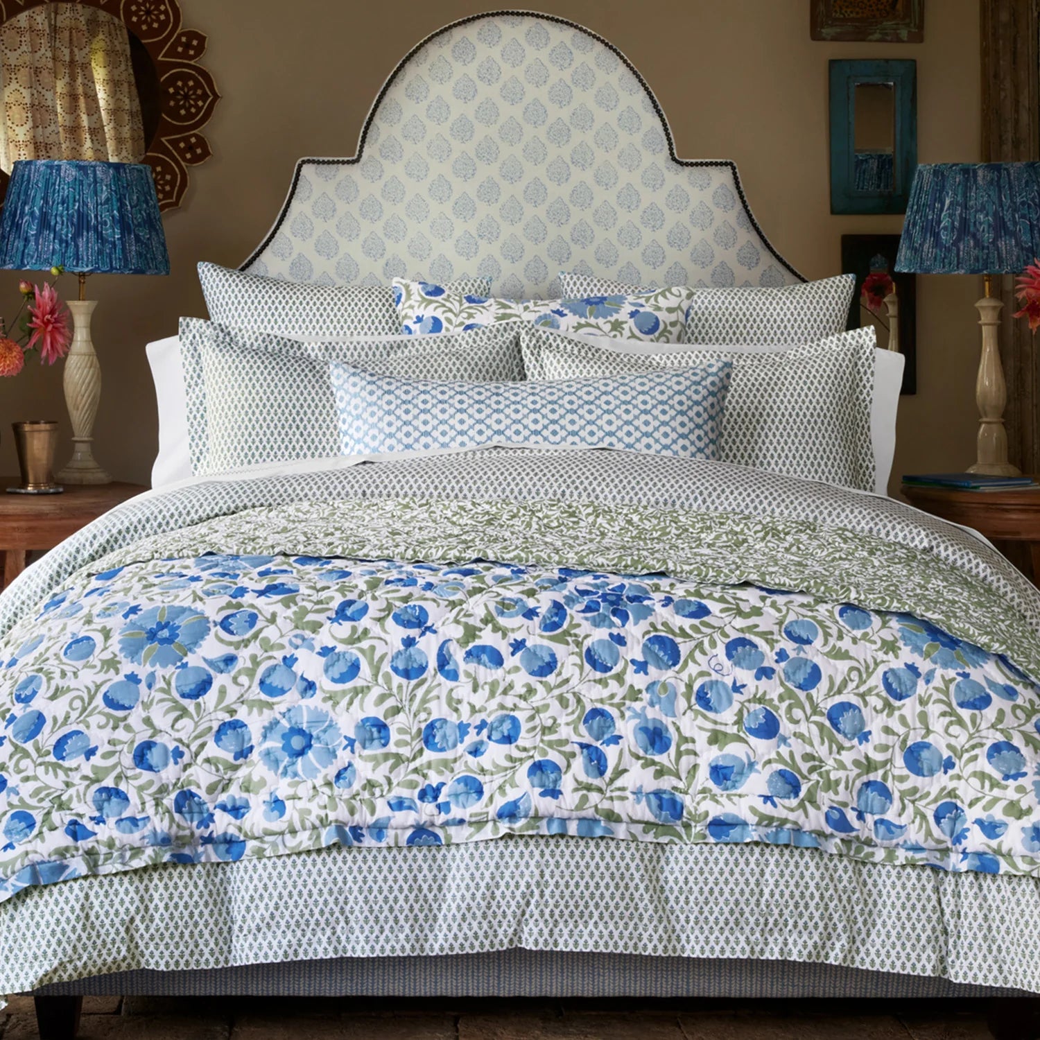 A neatly made bed features John Robshaw's Cala Duvet, a blockprint bouquet headboard, blue and white floral bedding, decorative pillows, and wooden nightstands with lamps and flowers for a cozy, garden-inspired room in soothing greens and blues.