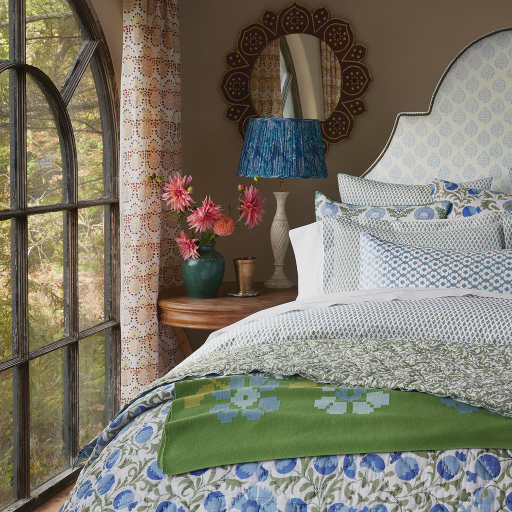 A cozy bedroom features a John Robshaw Cala Duvet in greens and blues, a green floral blanket, a side table with a vase of pink blockprint bouquet flowers, a blue lamp, decorative mirror, and an arched window with patterned curtains.