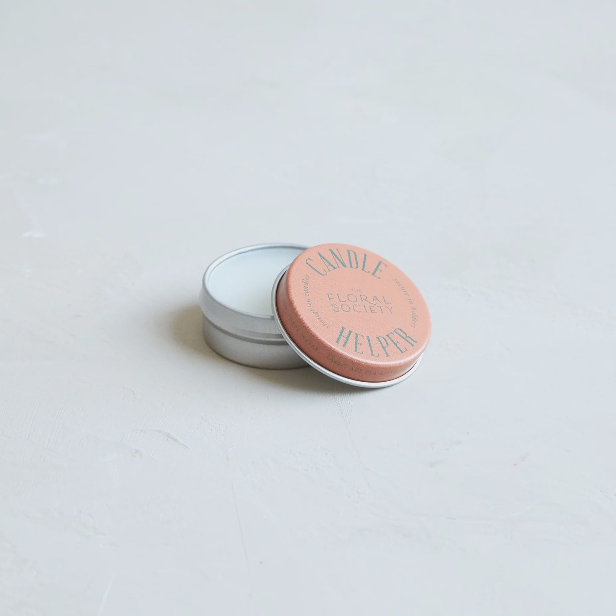 A small tin with a pink lid labeled "Candle Helper Wax" from The Floral Society rests on a smooth, light gray surface. The lid is slightly ajar, revealing the white candle wax inside that helps secure candles in place.