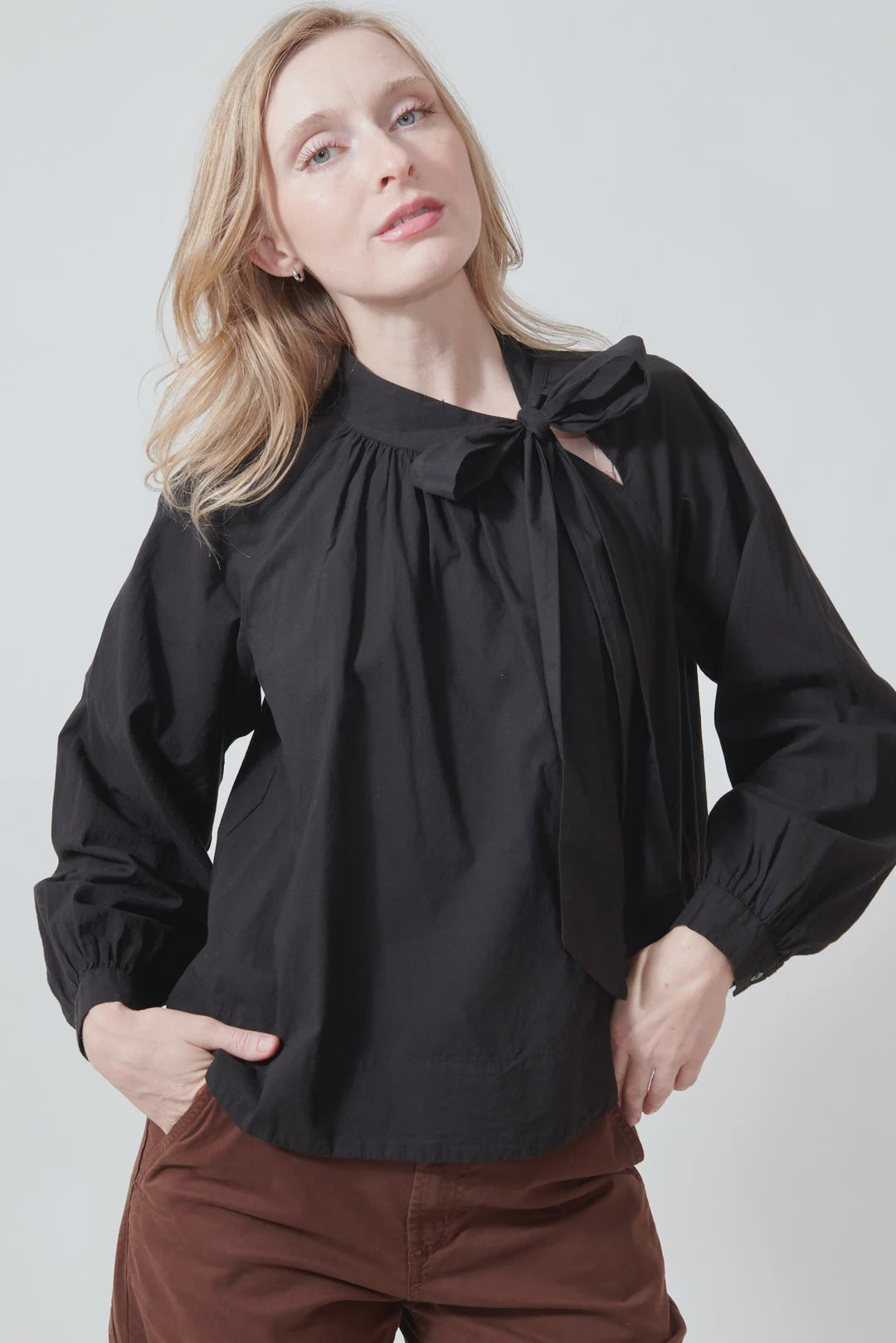 A person with long blonde hair wears the Xirena Leona Top, a black blouse with a bow tie neck detail, paired with brown pants and standing against a plain light background with one hand in their pocket.