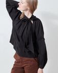A woman poses with one arm raised and her head turned to the side, wearing the Xirena Leona Top—a black long-sleeve blouse in lightweight cotton poplin with a feminine bow tie—paired with brown pants against a plain light background.
