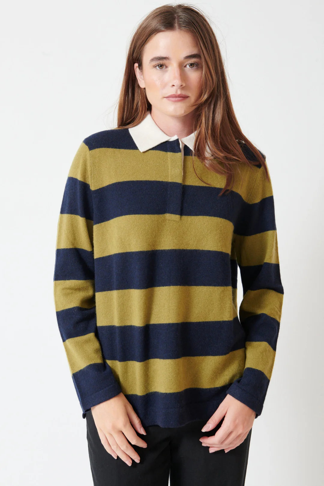 A person with long brown hair wears the White + Warren Cashmere Rugby Stripe Polo, featuring wide olive green and navy blue stripes, paired with black pants, standing against a plain white background.