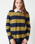 A person with long brown hair wears the White + Warren Cashmere Rugby Stripe Polo, featuring wide olive green and navy blue stripes, paired with black pants, standing against a plain white background.