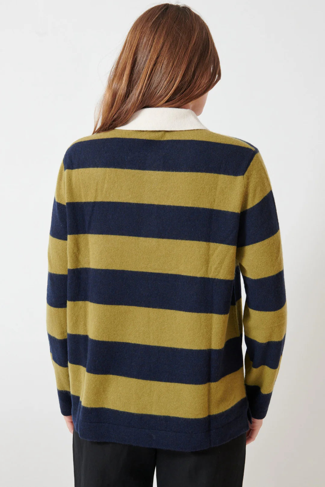 A person with long brown hair, seen from behind, wears the White + Warren Cashmere Rugby Stripe Polo—a navy blue and olive green striped sweater with a white collar—paired with black pants.