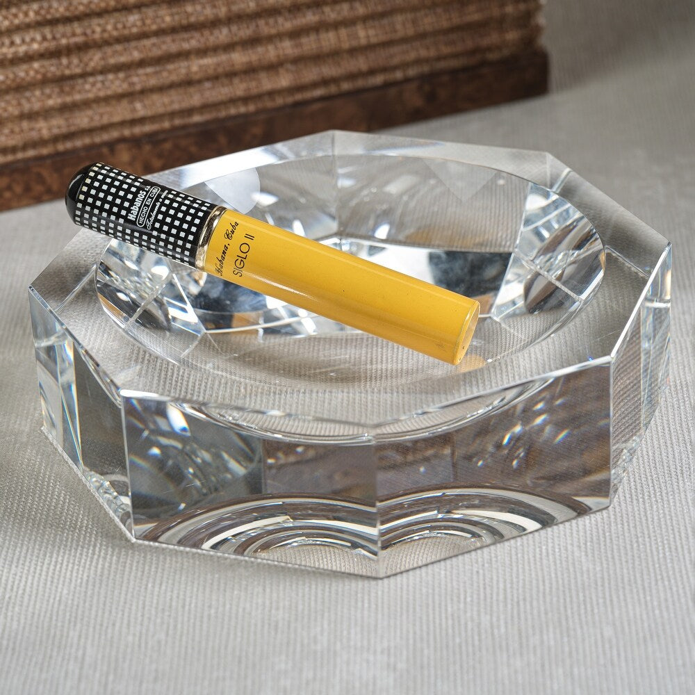 A single yellow cigar in a clear glass tube rests on the Zodax Octagon Bowl, a faceted crystal-like glass ashtray set on a beige textured surface, with brown woven material in the background for a modern look.