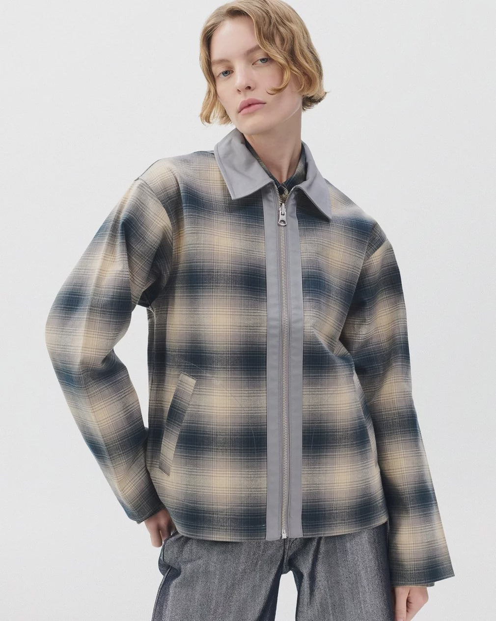 A person with short, wavy blonde hair models the Rag & Bone carrie reversible jacket in muted beige, blue, and gray plaid tones, paired with gray pants, against a plain white background.