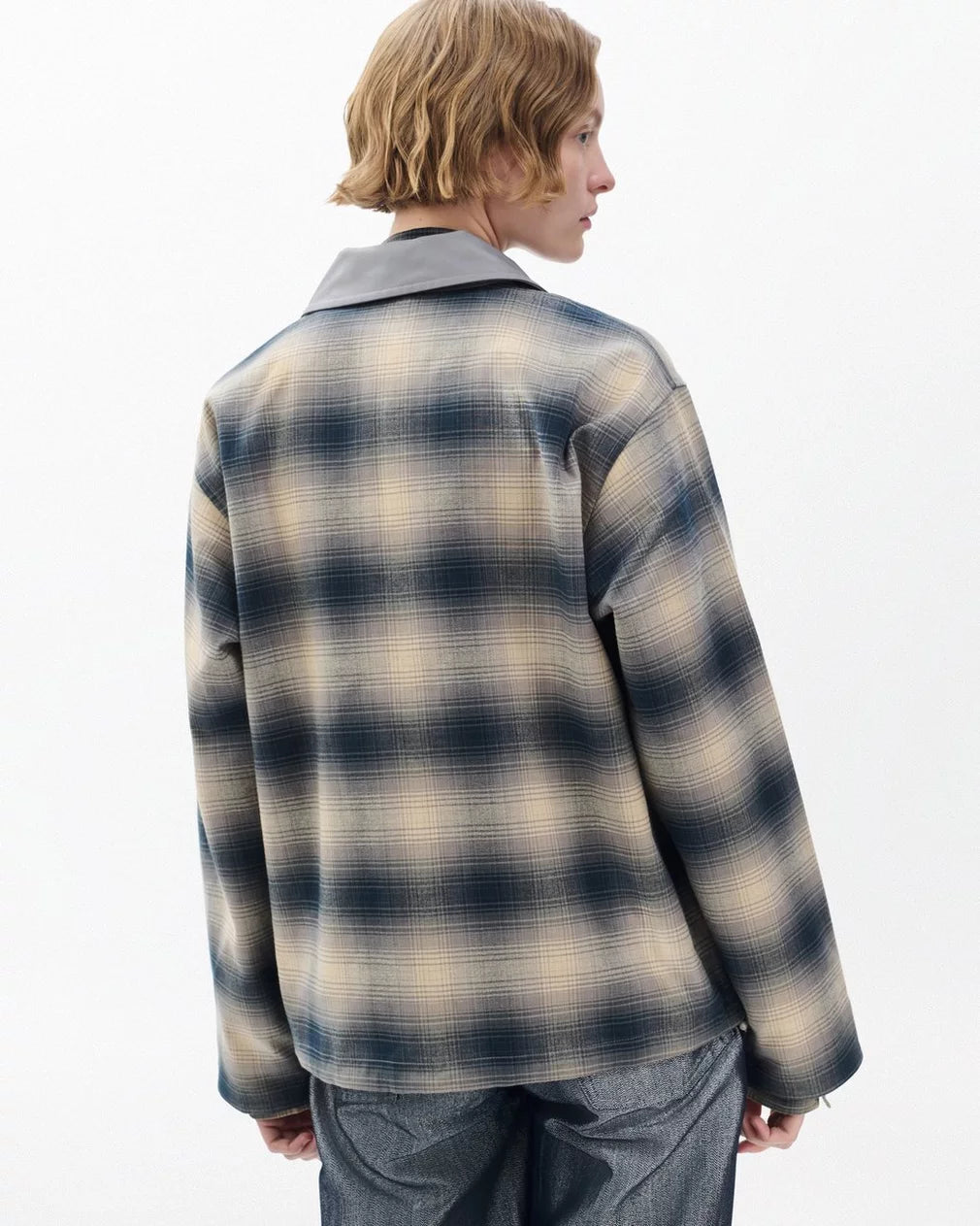 A person with short, wavy blond hair stands with their back to the camera, wearing the Rag & Bone carrie reversible jacket in beige and navy plaid with a gray collar and gray pants against a plain light background.