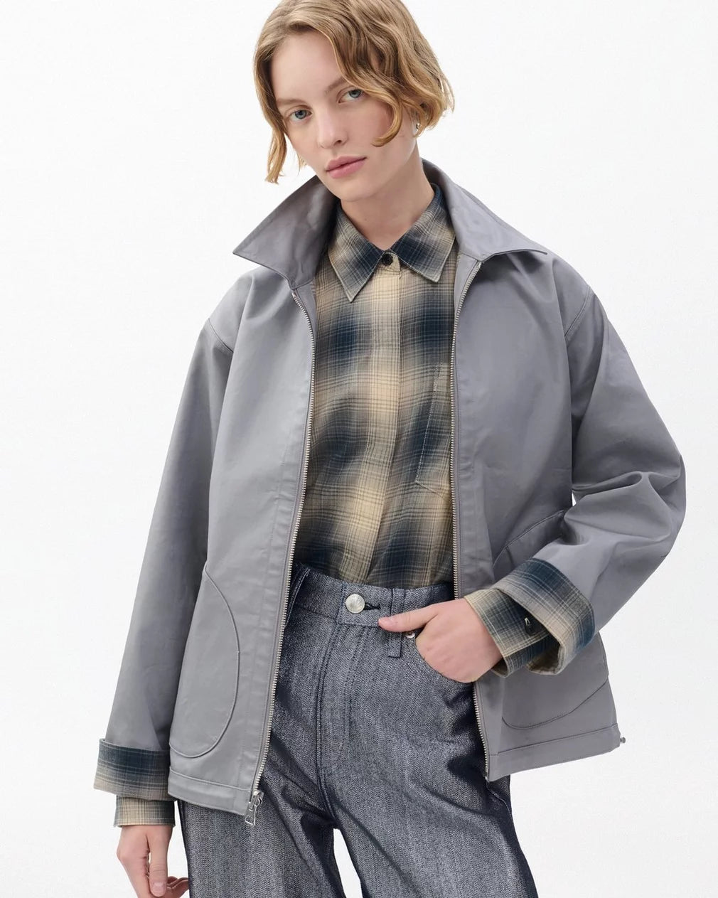 A person with short, wavy blonde hair wears the Rag & Bone carrie reversible jacket over a plaid shirt and high-waisted gray trousers, posing with one hand in their pocket against a plain white background.