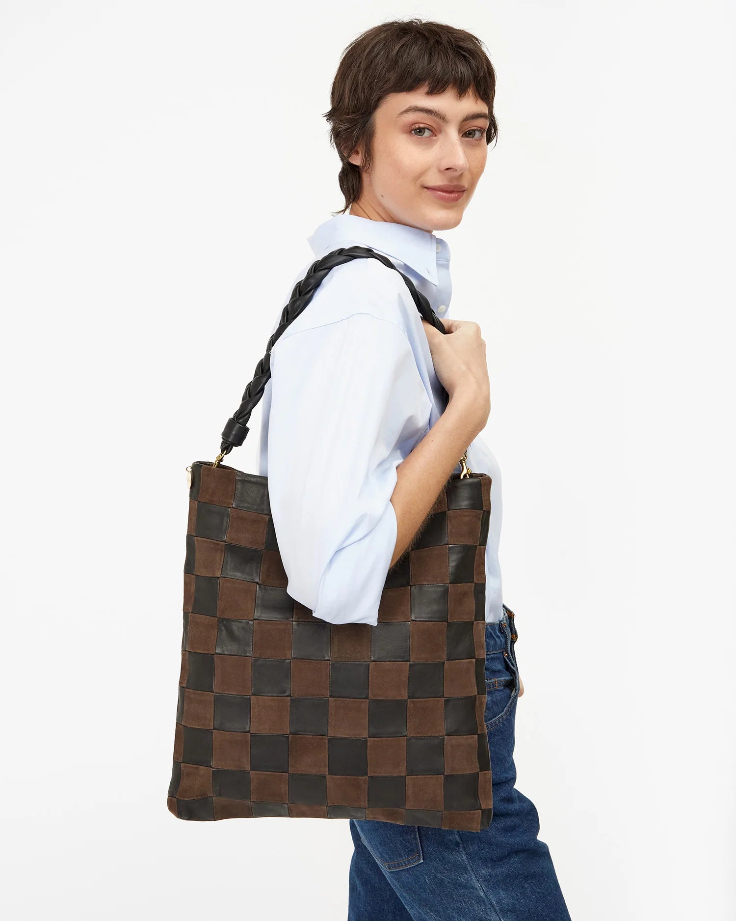 A person with short brown hair, in a light blue shirt and jeans, holds a Clare Vivier Carryall Clutch w/ Tabs featuring brown and black checks, braided strap, chambray lining, and interior zip pocket. Background is plain white.
