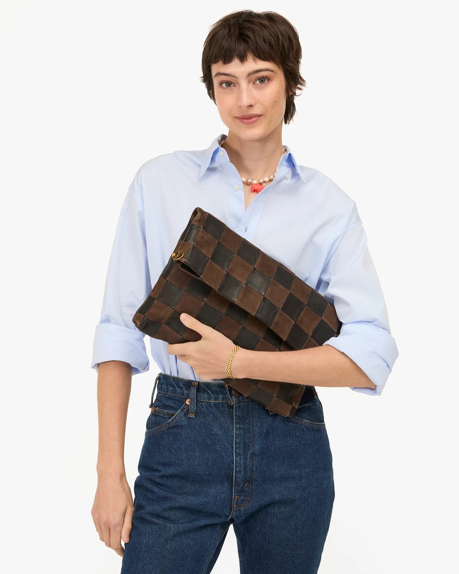 A person with short brown hair, dressed in a light blue shirt and blue jeans, holds the Clare Vivier Carryall Clutch w/ Tabs—a large brown woven bag with a magnetic closure—against a plain white background.