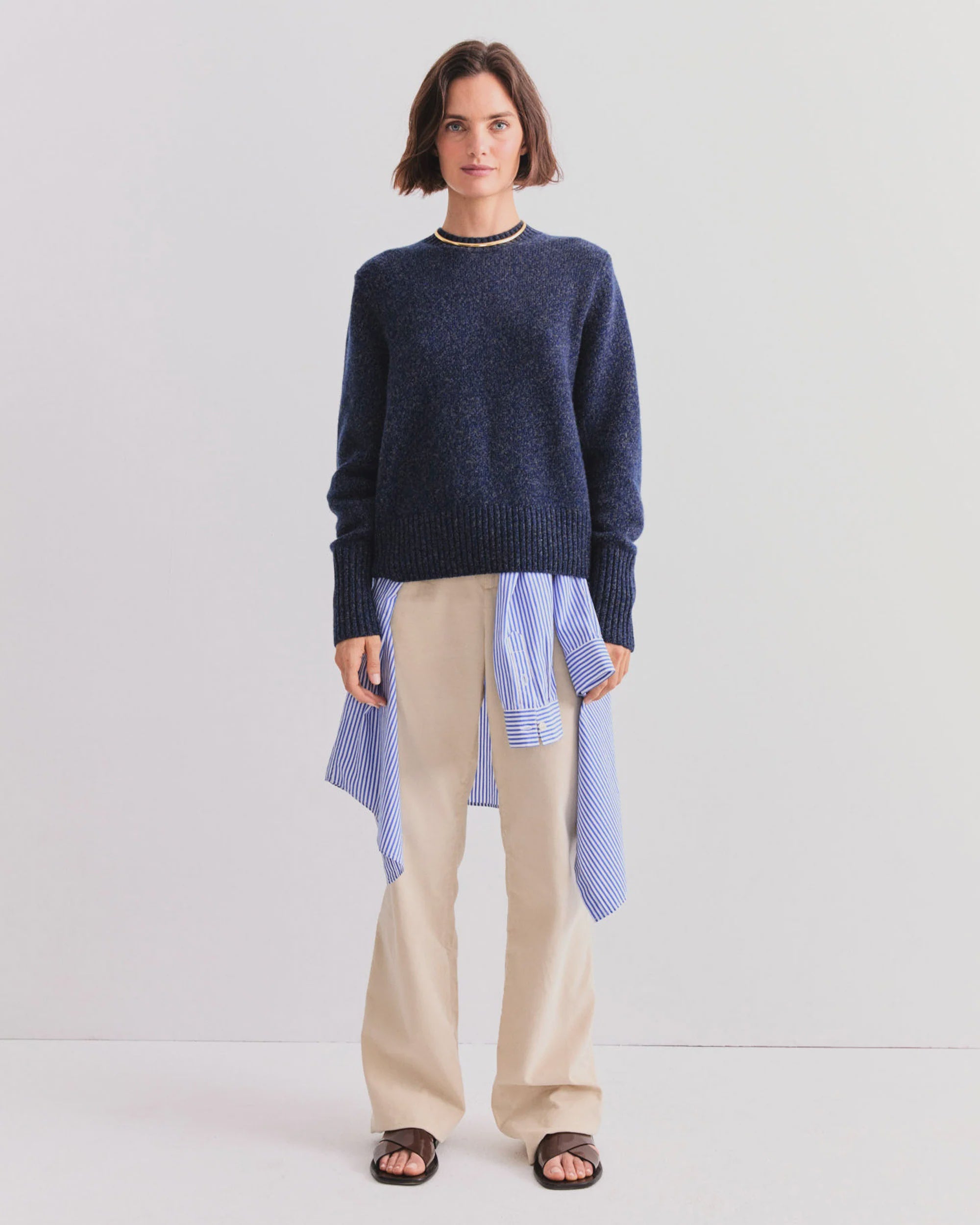 A woman leans against a white wall wearing Kule’s The Cashmere Tatum crewneck in navy, layered over a blue-striped shirt, with beige pants, black sandals, and a simple necklace—an effortless wardrobe staple.